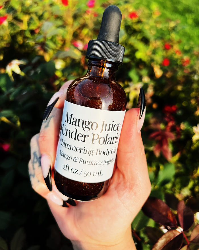 A hand with black and white manicured nails holds an amber glass dropper bottle of Mango Juice Under Polaris Shimmering Body Oil against a blurred background of green foliage and red flowers.