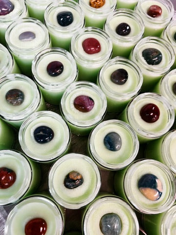 A close-up, top-down view of multiple small clear glass jars filled with pale green body butter, each topped with a smooth polished ocean jasper stone in colors like red, blue, black, and gray.