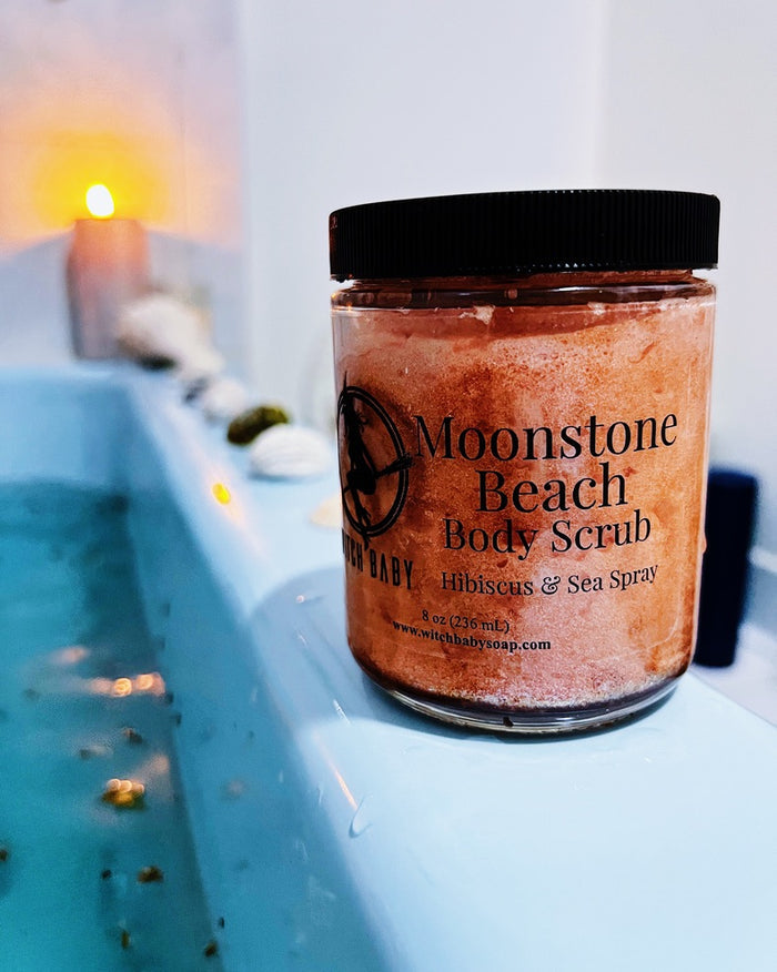 A jar of Moonstone Beach Body Scrub by Witch Baby Soap sits on the edge of a blue bath tub with a lit candle glowing in the background.
