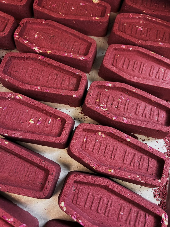 A close-up of multiple red coffin-shaped bath bombs arranged on a light surface. Each bath bomb is embossed with the words WITCH BABY