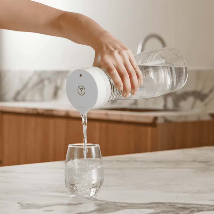 Person pouring water from a clear bottle with a white cap into a glass on a marble countertop.