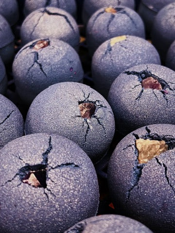 A close-up of several dark lavender, spherical bath bombs with a textured, amethyst and visible cracks, filling the frame.