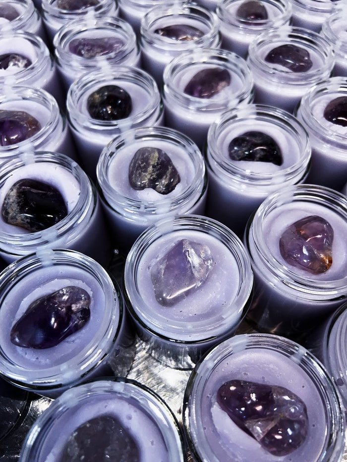 Multiple small clear glass jars filled with pale lavender-purple cream each topped with a polished dark purple amethyst crystal, arranged closely together.