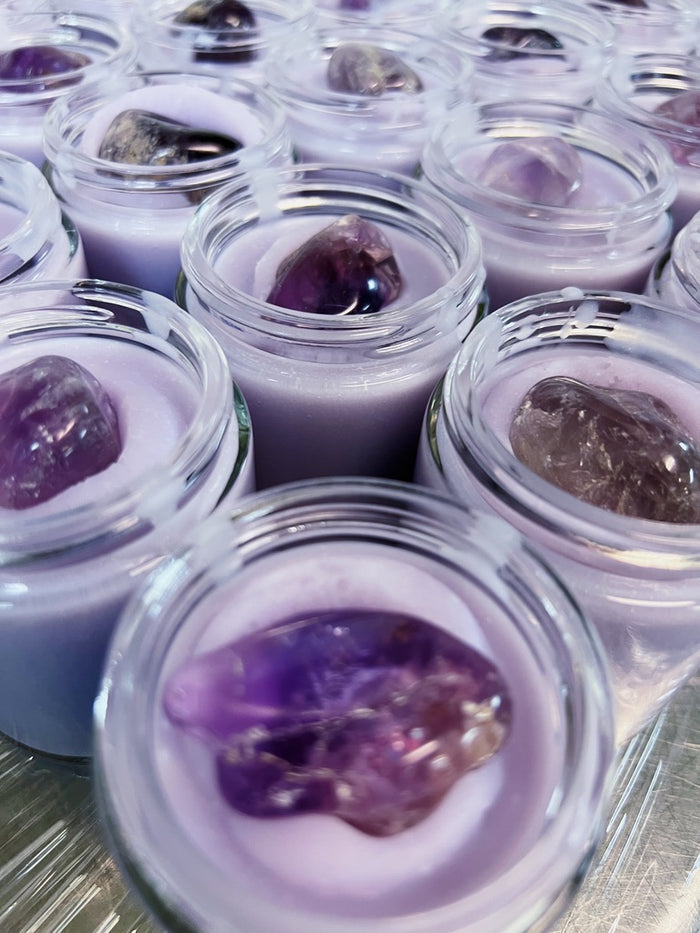 Multiple small clear glass jars filled with pale lavender-purple cream each topped with a polished dark purple amethyst crystal, arranged closely together.