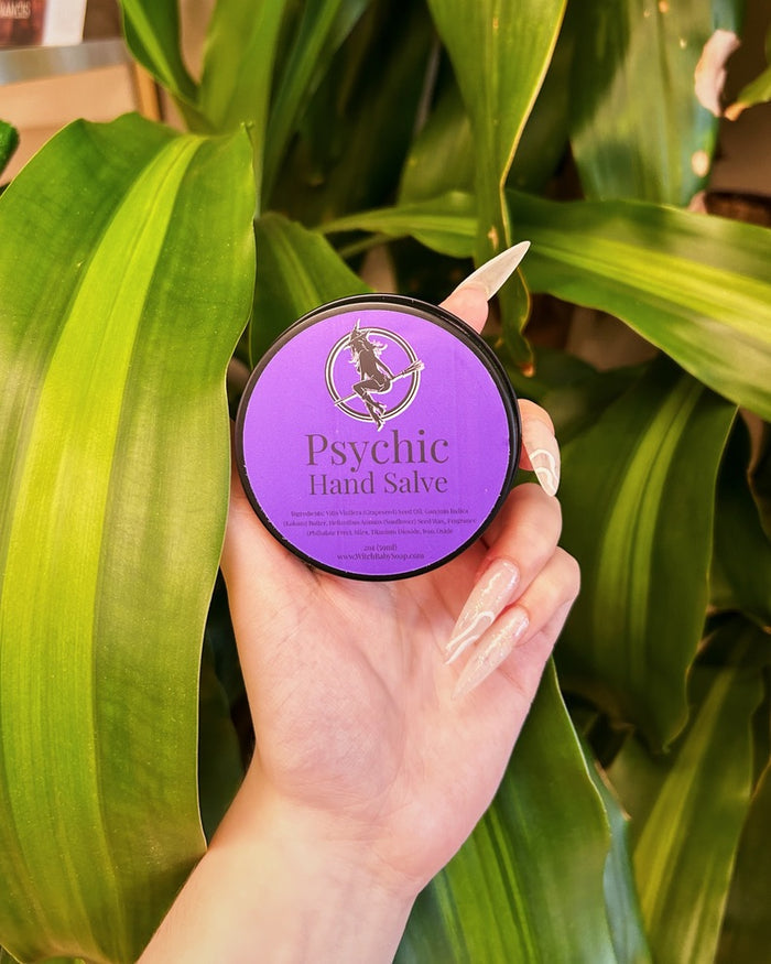 A hand holding a round black container of Psychic Hand Salve with a purple label, set against a background of large green houseplant leaves.