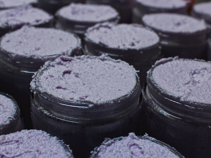 Close-up of several open dark glass jars filled with a textured lavender-purple scrub, arranged closely together.