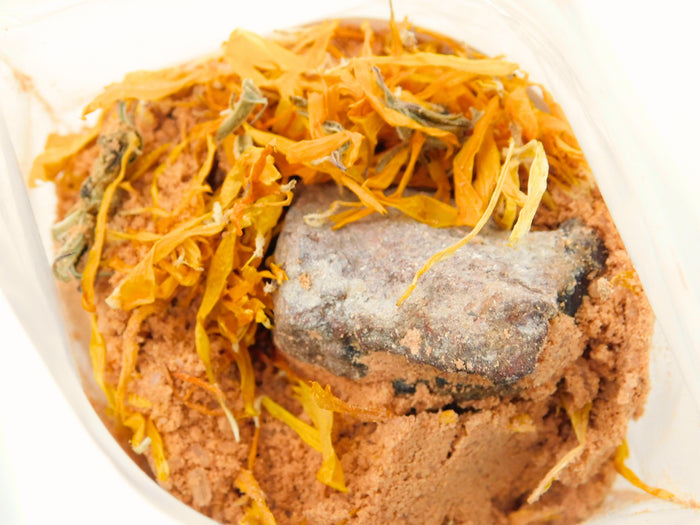 A close-up of a loose bath potion inside a clear biodebgradable plastic bag. The blend consists of a light orange powder base topped with dried orange-yellow calendula flower petals and a chunk of raw fire agate.