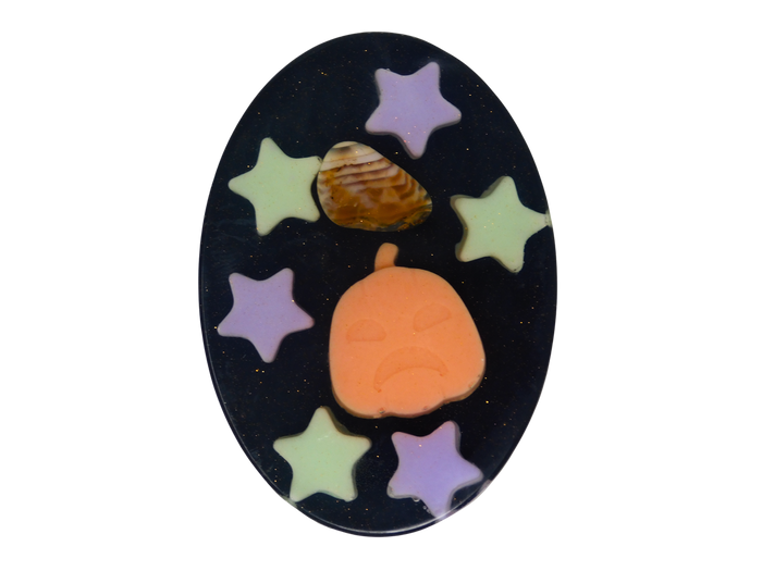 An oval black soap featuring a Halloween design with a carved orange pumpkin, a tumbled fire agate crystal, and six pastel star cutouts in purple and light green.