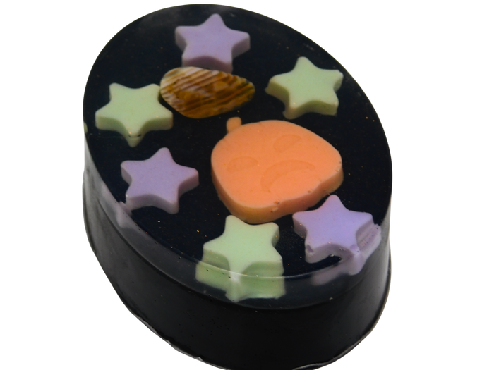 A round black soap with embedded pastel star shapes in purple and green, plus a small orange pumpkin-shaped piece in the center, and a tumbled fire agate crystal shown on a plain white background.