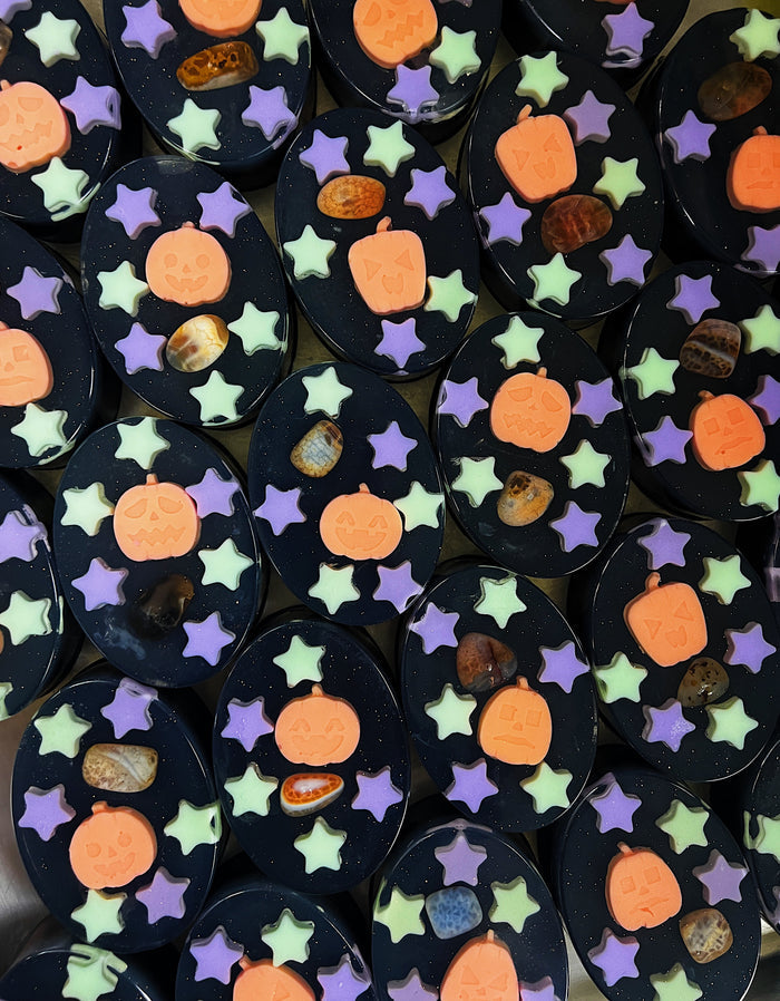 A close-up, top-down view of multiple Halloween-themed soap bars arranged side-by-side. The soaps are dark, featuring orange pumpkin shapes, light green star shapes, and purple star shapes, with small decorative stones embedded in the designs.