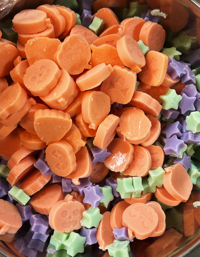 A close-up view of colorful Halloween soap embeds in a clear bowl, featuring many orange pumpkin pieces on top with purple and light green star-shapes underneath.