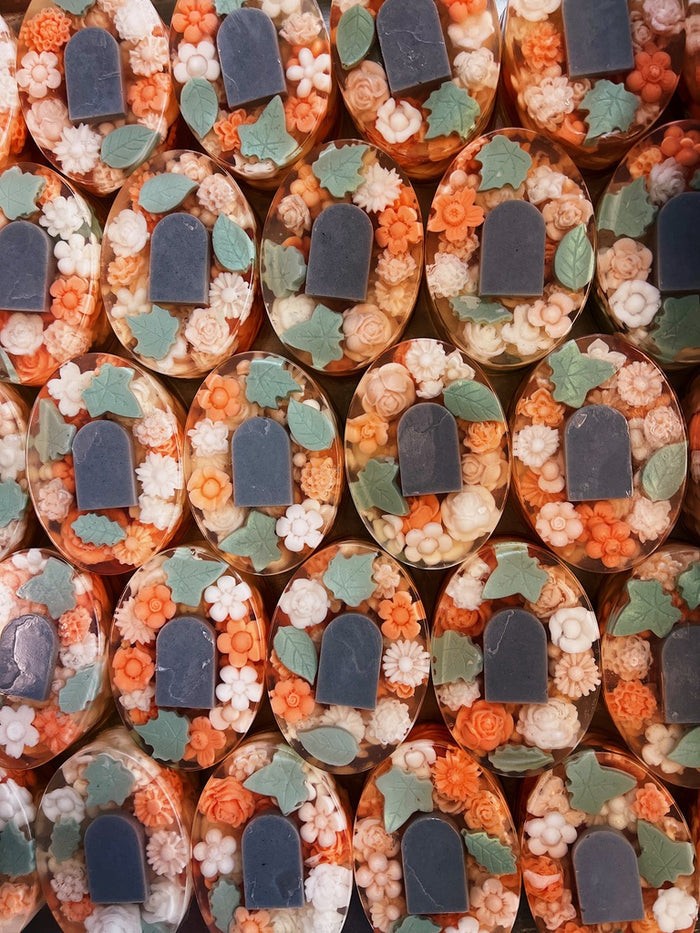 A close-up, top-down view of multiple oval-shaped soap bars arranged in a grid. Each soap is clear and filled with orange and cream flower-shaped pieces, green leaves, and a dark gray tombstone motif in the center.