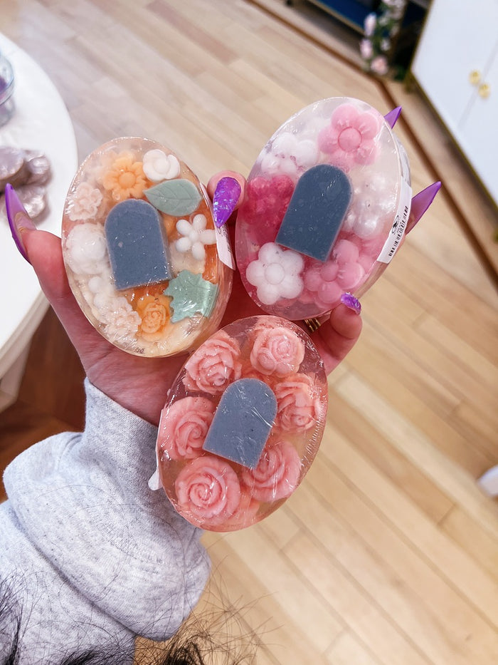 Three decorative translucent soaps held in a hand, featuring floral rosettes and small tombstone shapes in pink, peach, orange, white, and gray, against a light wood floor background.