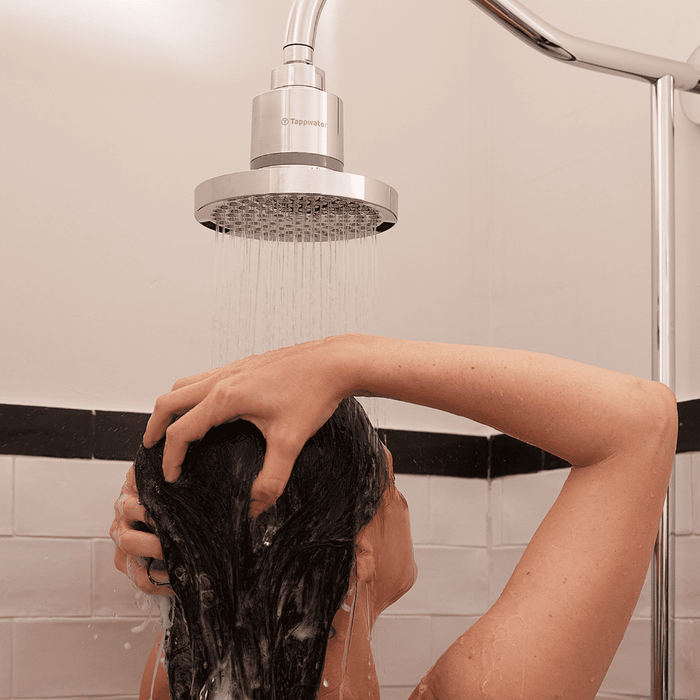 A person with dark hair washes their hair under the Tappwater ShowerPro Rain, a modern silver showerhead, as water cascades over their head and hands in a tiled shower. Their back faces the camera.