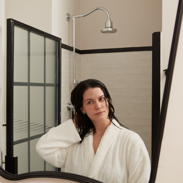 A woman in a white bathrobe with wet hair stands in a bathroom, gazing into the mirror. Behind her, the Tappwater ShowerPro Rain shower with elegant white tiles and a glass door is visible.