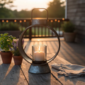 Small workman's lantern on an outdoor weathered wood porch table at dusk with potted herbs and linen napkin, warm golden candlelight