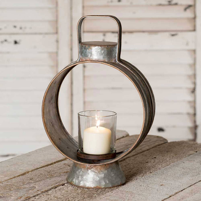 Small workman's lantern with circular galvanized metal frame, carry handle, and glass chimney holding a white pillar candle on a weathered wood surface
