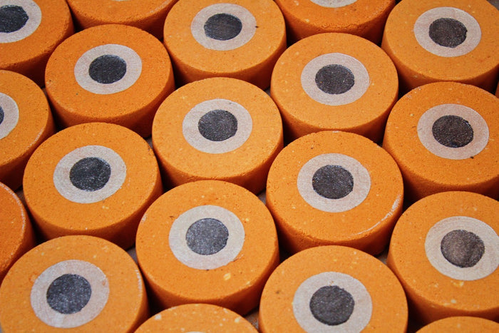 A close-up, top-down view of multiple orange cylindrical bath bombs arranged in a grid, each with a designed like an orange evil eye