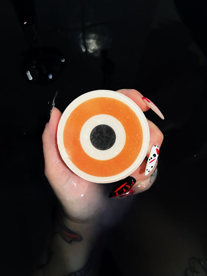 A hand with long decorated nails holds a round orange evil eye soap