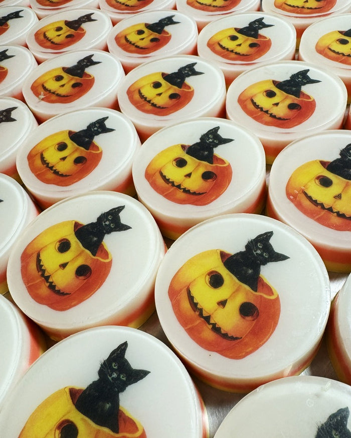 A close-up of multiple round white soaps stacked together, each decorated with a jack-o'-lantern face wearing a black witch hat, with a black cat sitting on the pumpkin.