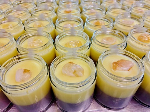 Multiple small clear glass jars filled with layered yellow and brown body butter, each topped with a citrine crystal, arranged closely together on a surface.