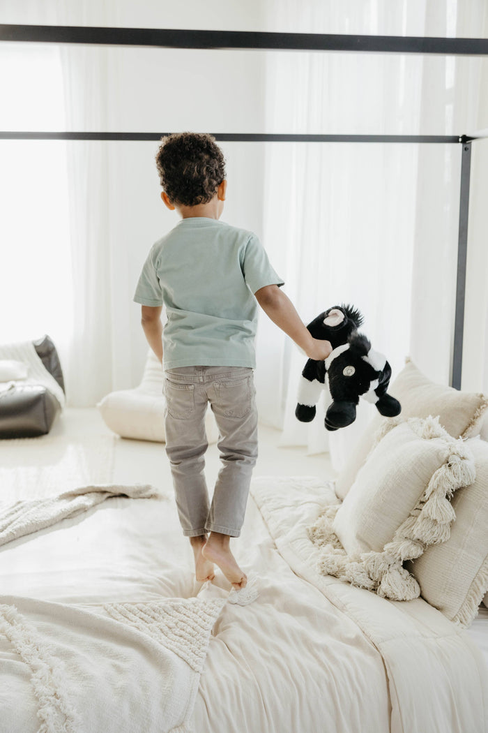 Young boy standing on a white bed holding a Wild Onez black and white cow plush by its leg, back facing camera