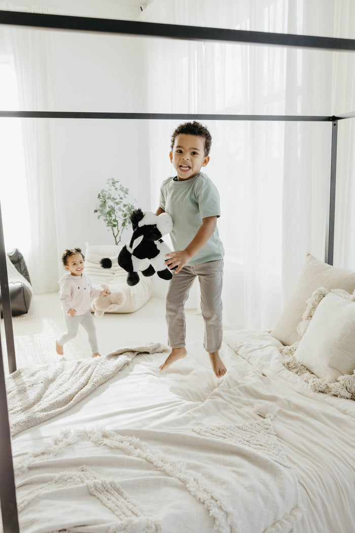 Smiling boy jumping on a white bed holding a Wild Onez black and white cow plush, younger girl playing in the background
