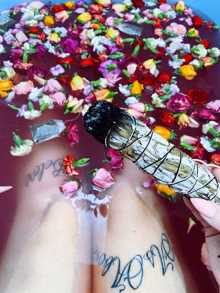 A hand holds a yerba santa smudge bundle with a dark, burned tip over a person's legs in a bath of pink water filled with colorful flower petals.